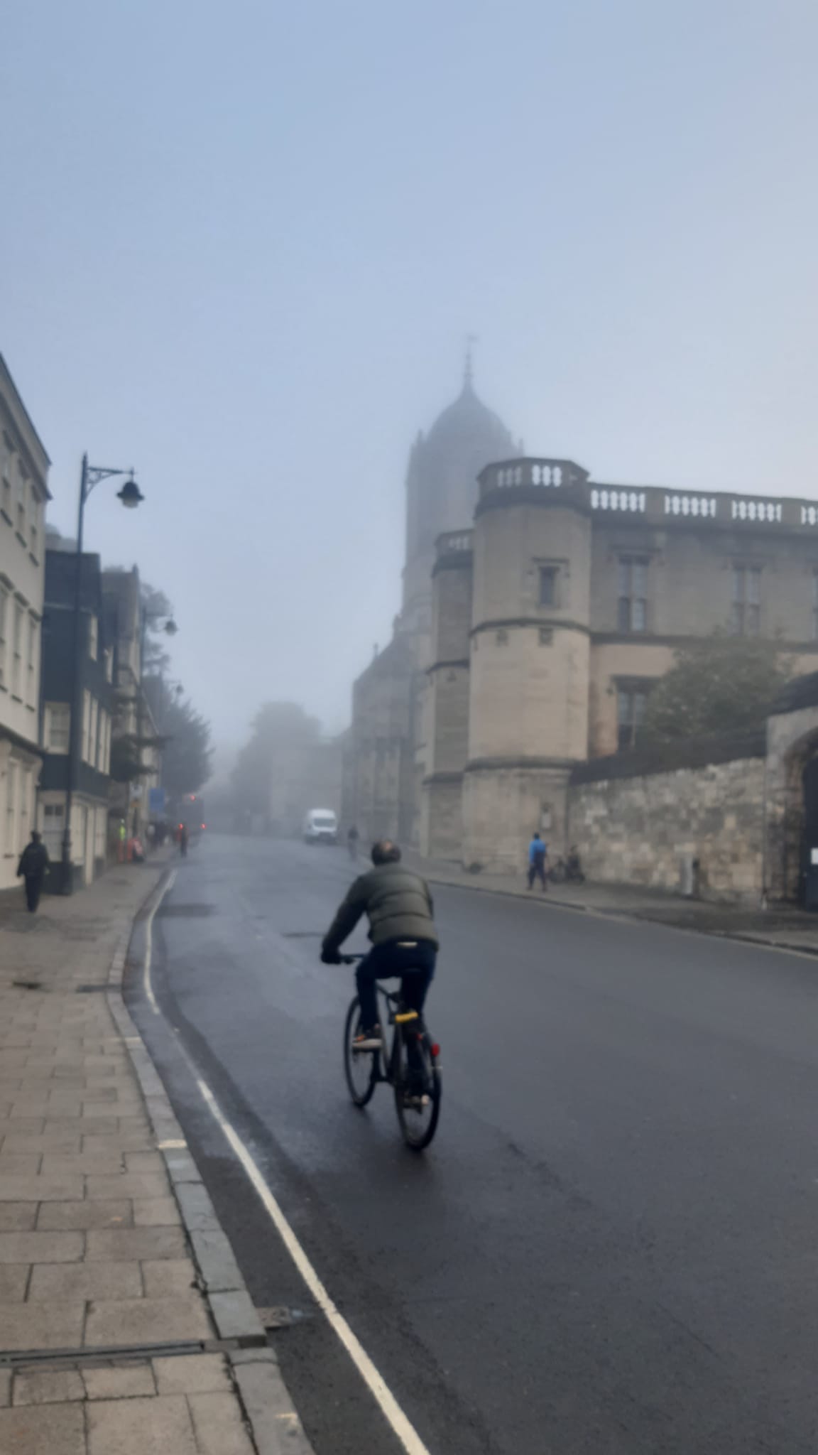 A view of St Aldates street on a misty grey morning with Christchurch College's Tom Tower appearing through the mist and a cyclist on the street ahead.